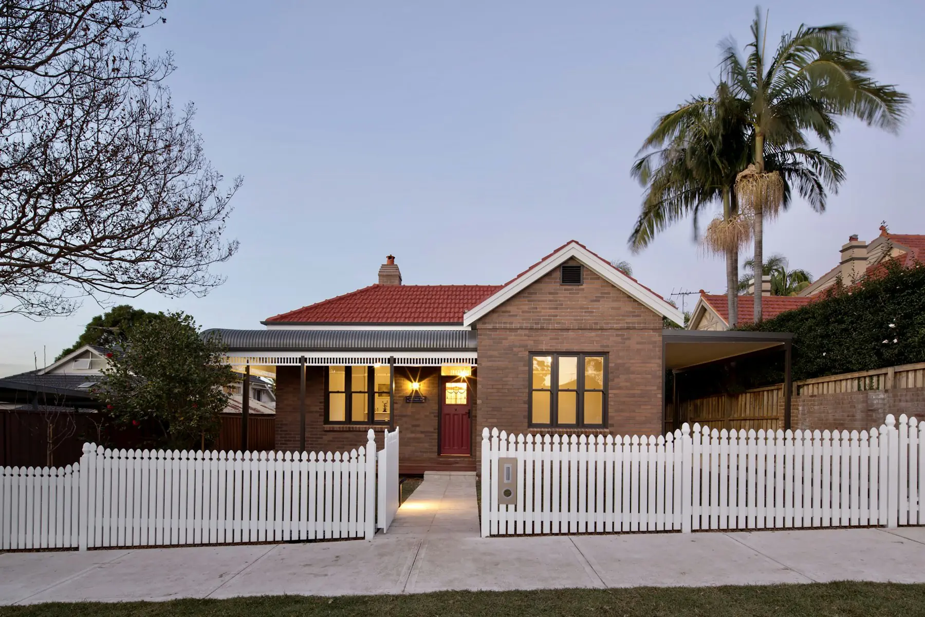A red-brick Australian home with a white picket fence at dusk